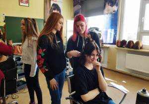 Dziewczynka siedzi na fotelu, dwie uczennice przygotowują się do wykonania fryzury.